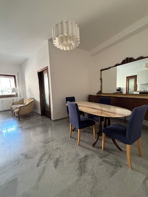 A dining area with a marble table, blue chairs, a hanging light fixture, and a view of a kitchen through a mirror. - New Listing (Roma)