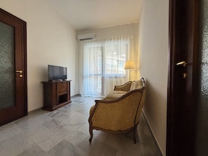 A room with a television on a wooden cabinet, a sofa, a lamp, and a door. - New Listing (Roma)