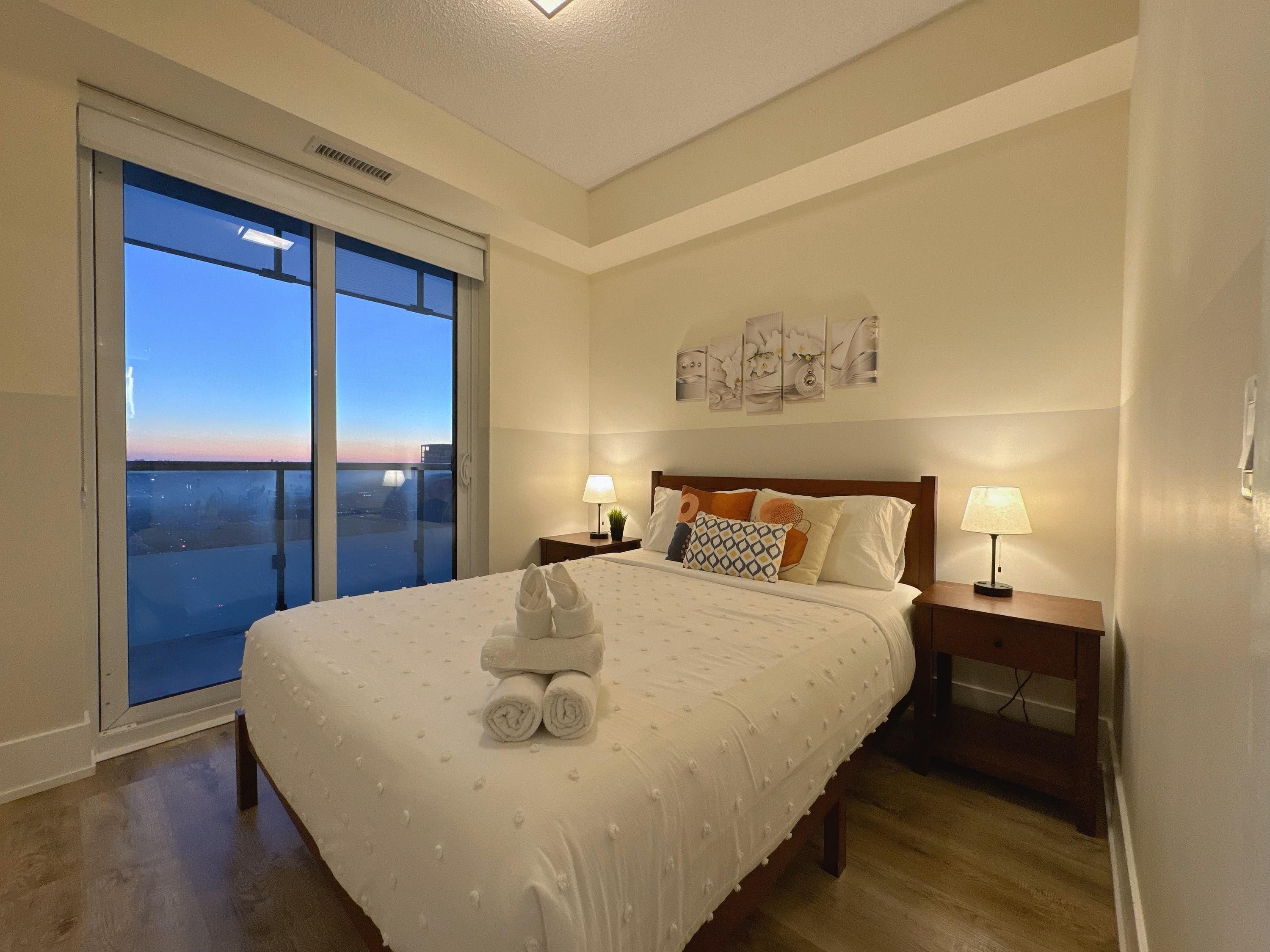 A hotel room with a large bed, two bedside tables with lamps, a view of the city through a large window, and artwork on the wall.