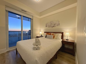 Quarto de hotel com uma cama grande, duas mesas de cabeceira com luminárias, vista da cidade por uma janela ampla e obras de arte na parede.