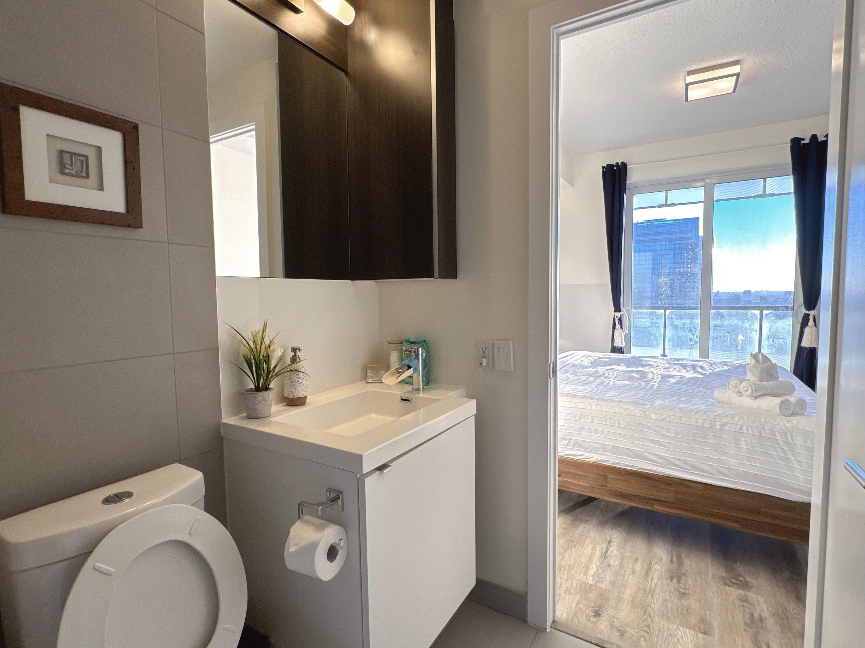 A modern bathroom with a toilet, sink, and mirror. A bed with a view of the city is visible through an open door.