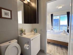 Uma casa de banho moderna com sanita, lavatório e espelho. Uma cama com vista para a cidade é visível através de uma porta aberta.