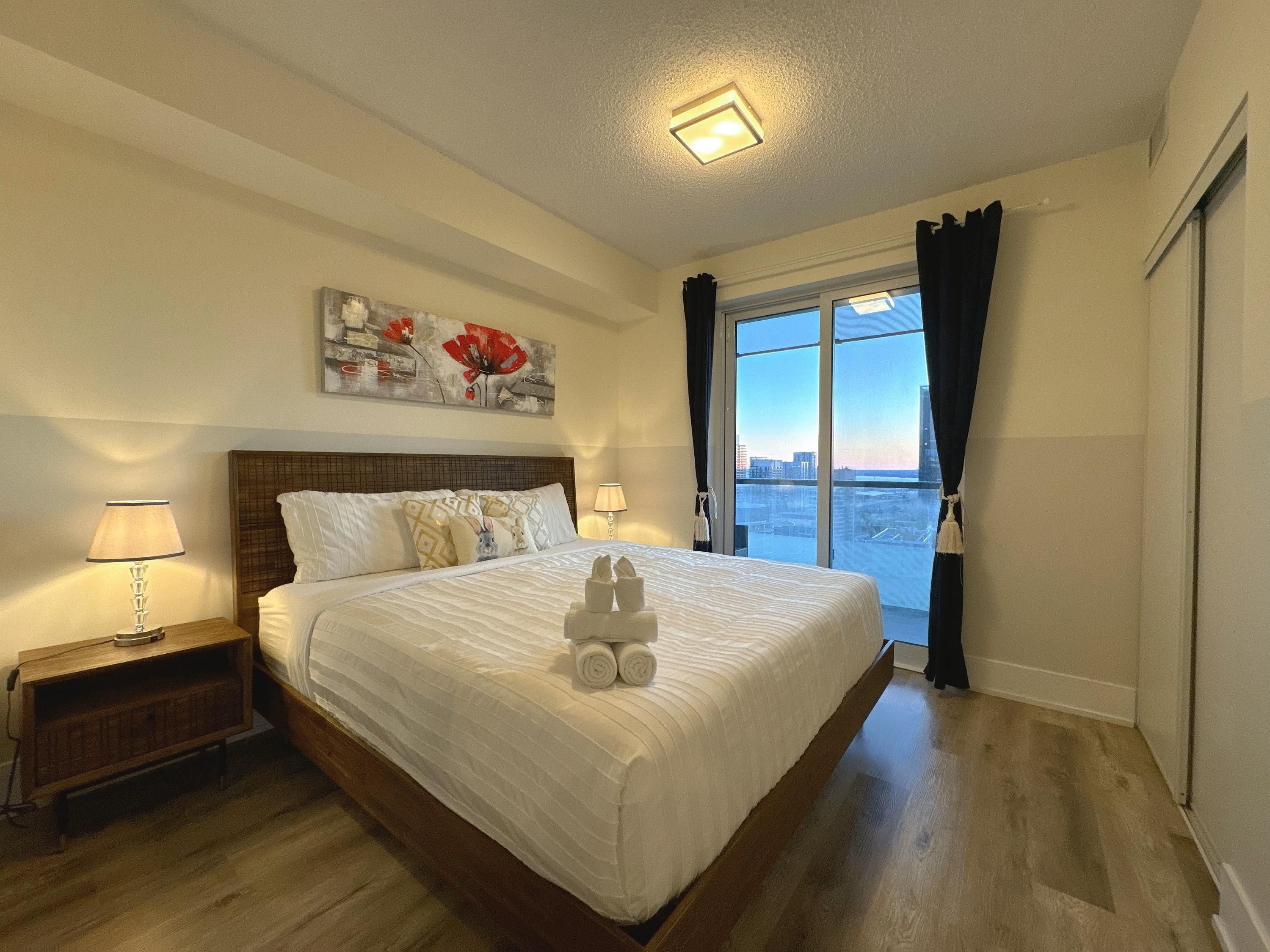 A hotel room with a large bed, a nightstand, a lamp, a painting, and a view of the city through a window.