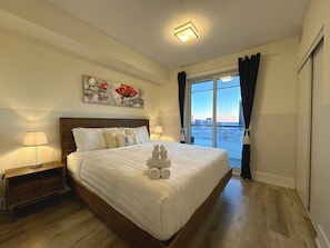 Quarto de hotel com uma cama grande, mesa de cabeceira, abajur, quadro e vista da cidade pela janela.