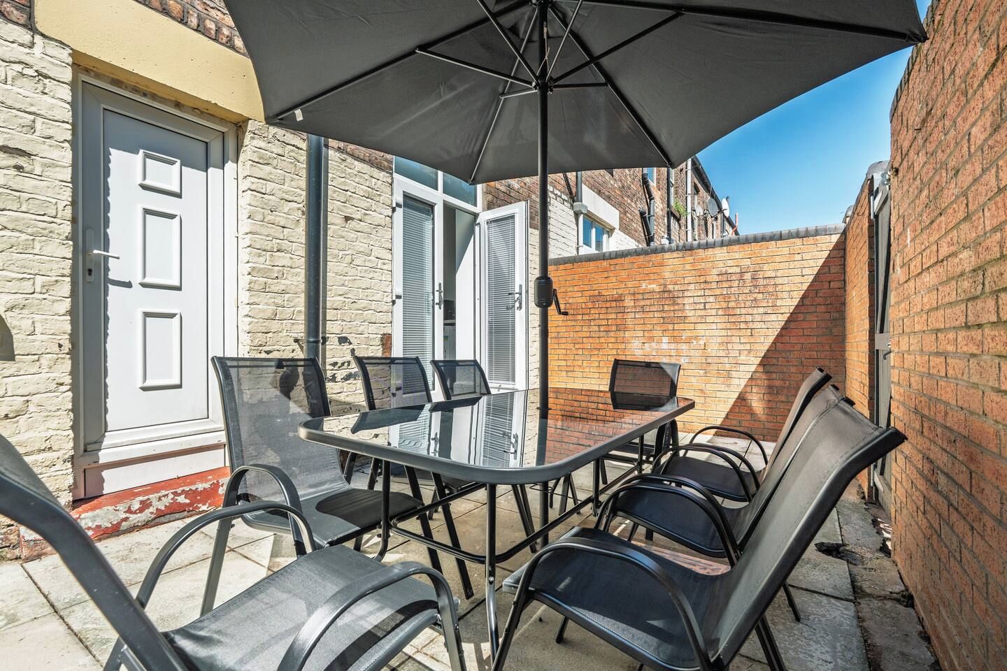 An outdoor patio with a glass table and chairs, a black umbrella, and a brick wall.