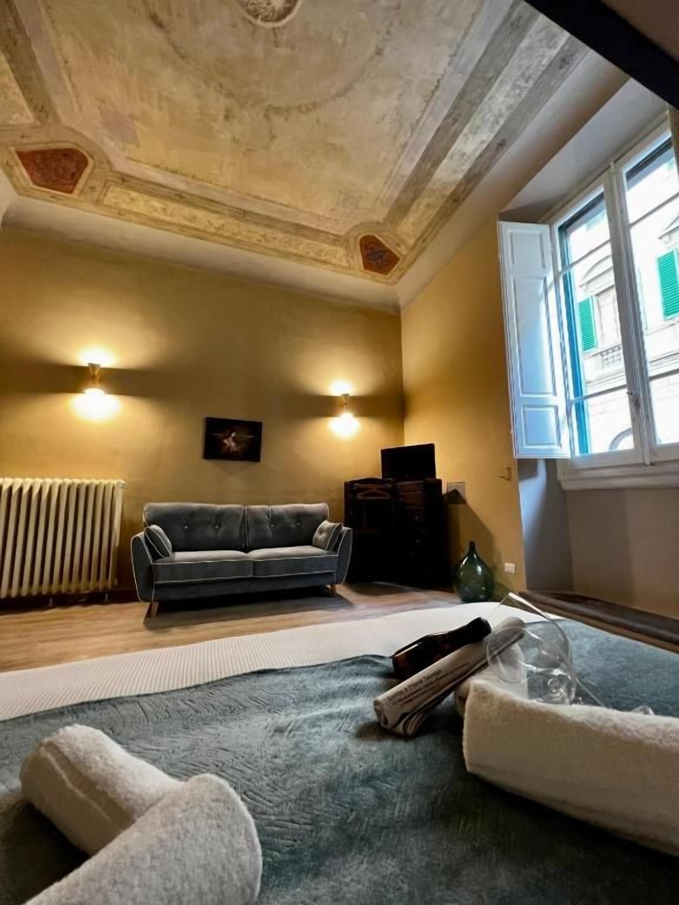 Une pièce comprenant un canapé, un radiateur, une fenêtre avec des volets et un plafond orné d’éléments décoratifs.
