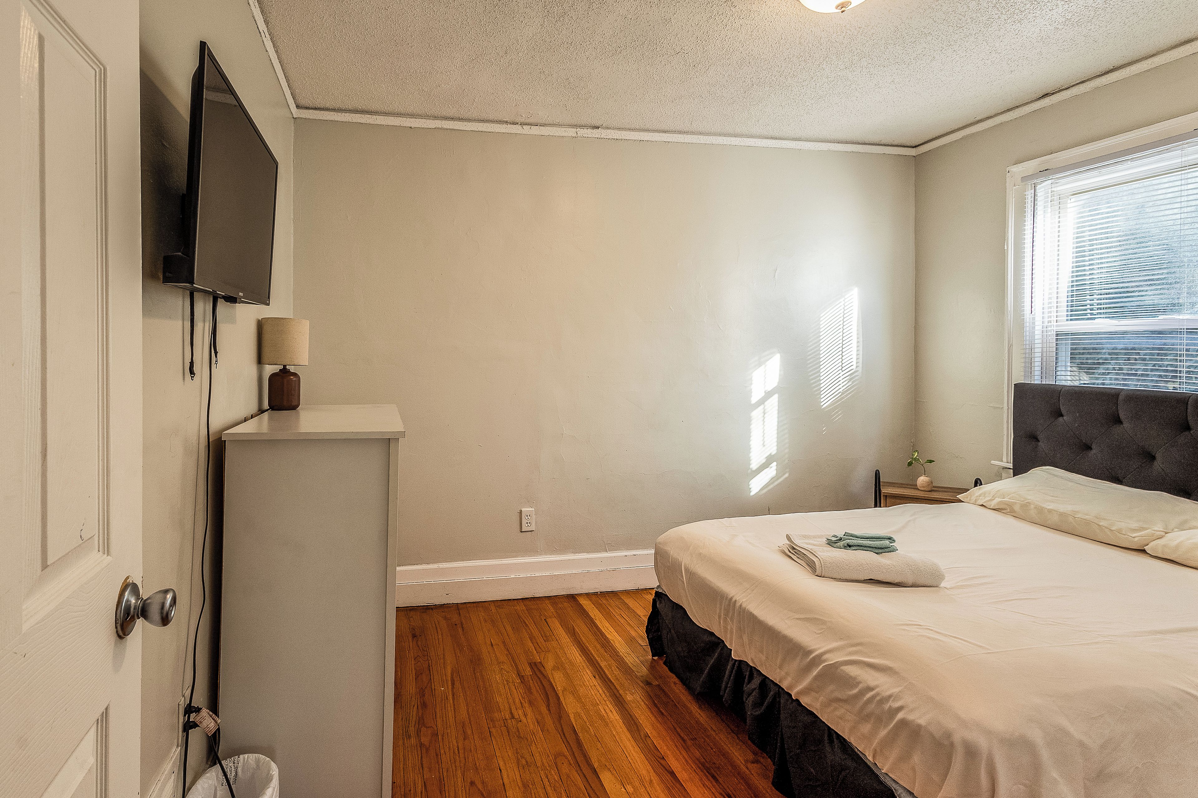 A bedroom with a bed, a television mounted on the wall, a bedside table with a lamp, and a window with blinds.