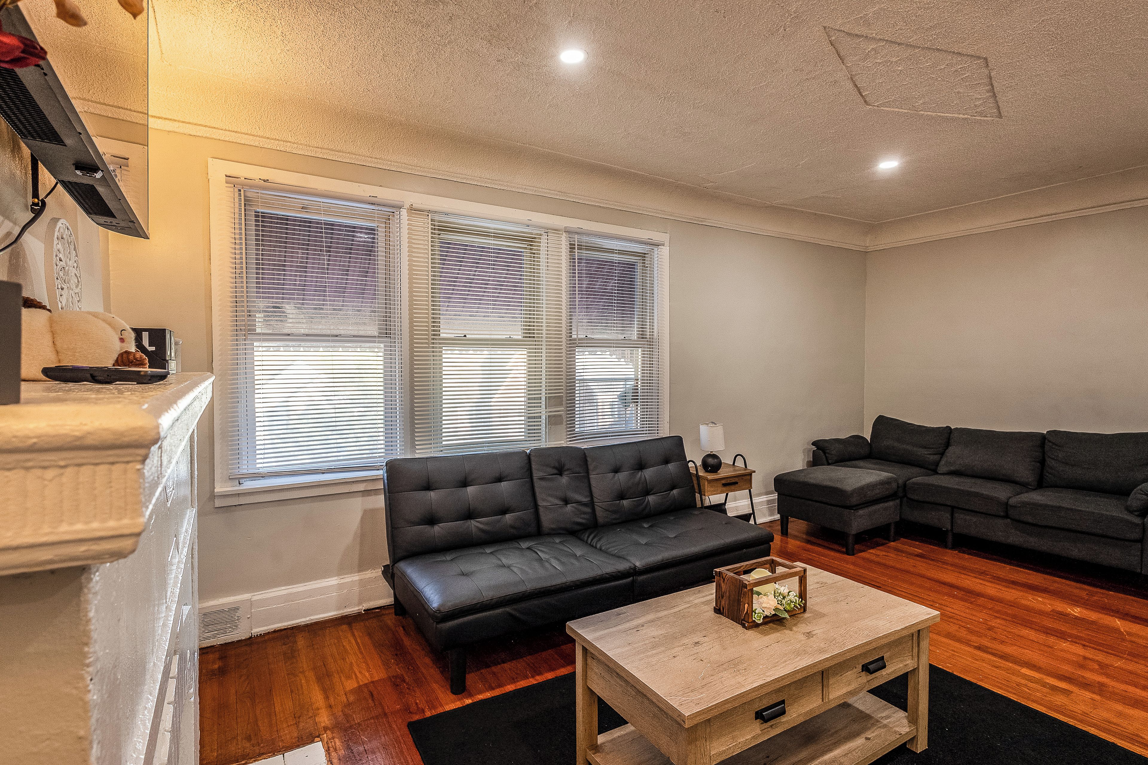 A living room with a black sofa, a wooden coffee table, and a window with blinds.
