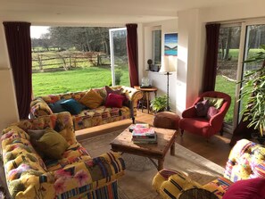 Ein Wohnzimmer mit Sofa, Sessel und Couchtisch, Blick auf eine grüne Landschaft durch große Fenster, und eine Lampe auf einem Beistelltisch.