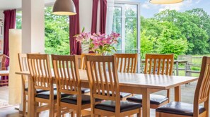 Un comedor con una mesa y sillas de madera, un florero con flores y vistas al verdor a través de amplias ventanas.