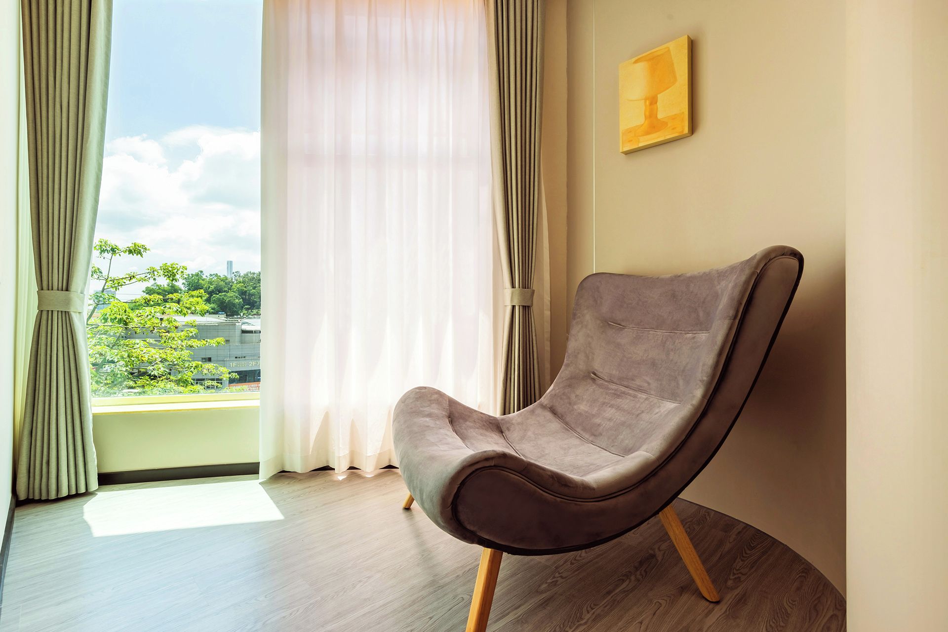 Uma cadeira moderna com pernas de madeira num quarto com uma janela grande, soalho de madeira e um quadro na parede.