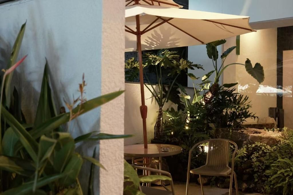 Un patio al aire libre con una mesa redonda y sillas, rodeado de plantas y un paraguas grande que da sombra.