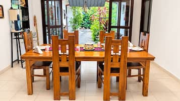 A dining area with a wooden table and chairs, a view of greenery outside, and a shelf with various items on the left.