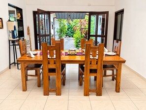 Un área de comedor con una mesa y sillas de madera, vista a zonas verdes exteriores y una estantería con varios objetos a la izquierda.