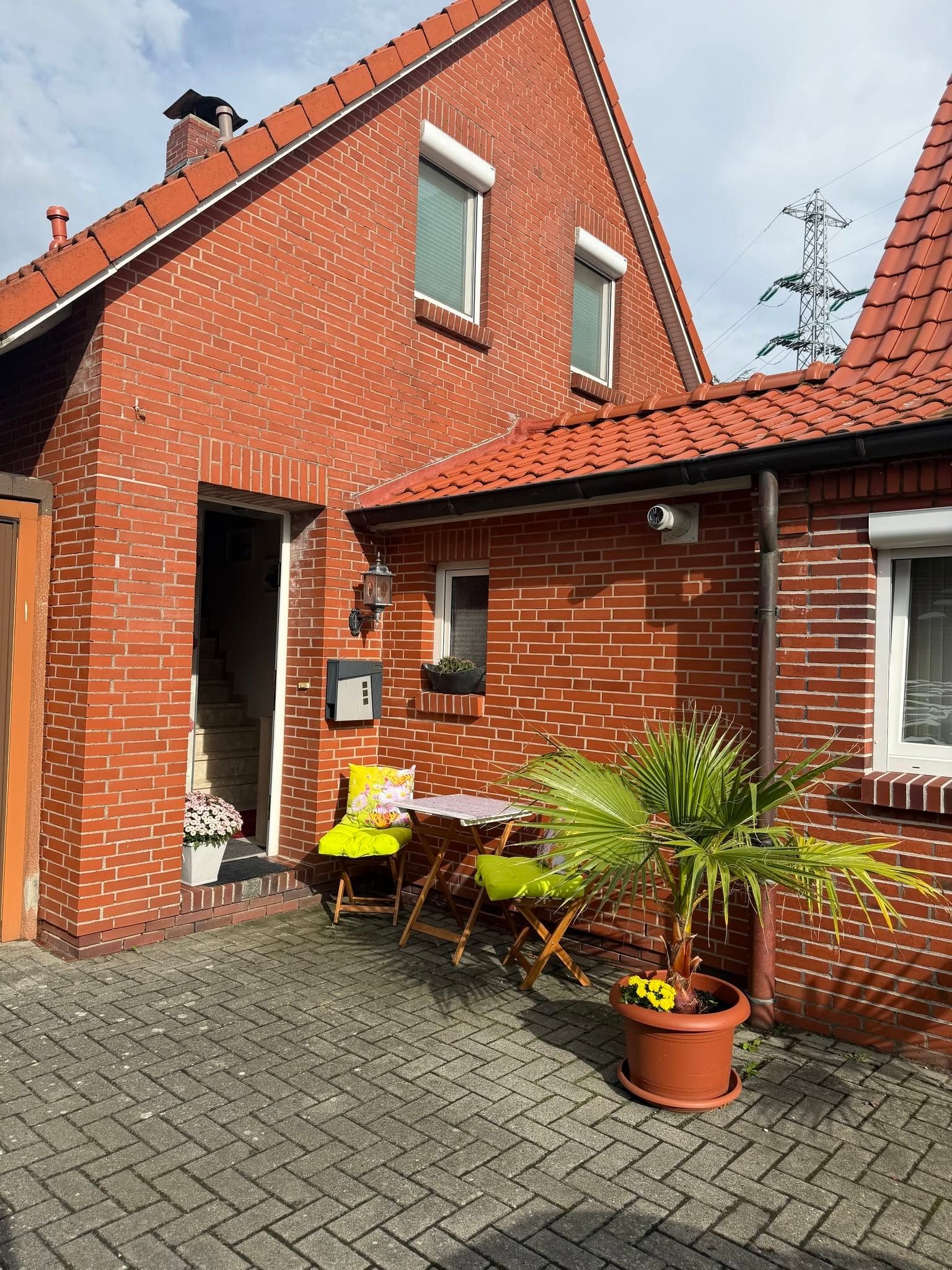 Ein Backsteinhaus mit Ziegeldach, eine kleine Terrasse mit Tisch und Stühlen sowie Kübelpflanzen.