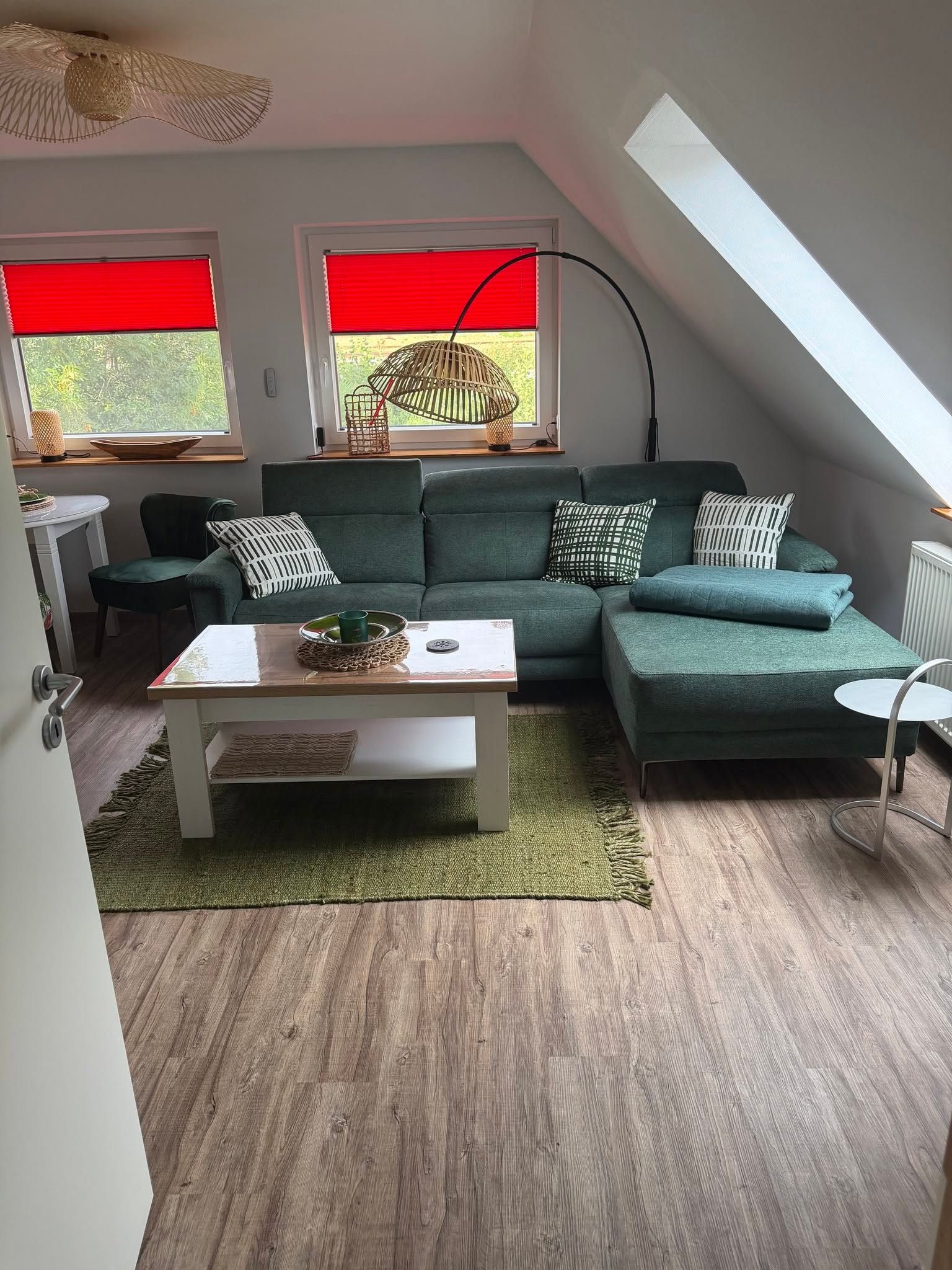 Ein modernes Wohnzimmer mit einem grünen Ecksofa, einem weißen Couchtisch und einem grünen Teppich. Es gibt zwei Fenster mit roten Jalousien, einen Deckenventilator und ein Oberlicht.