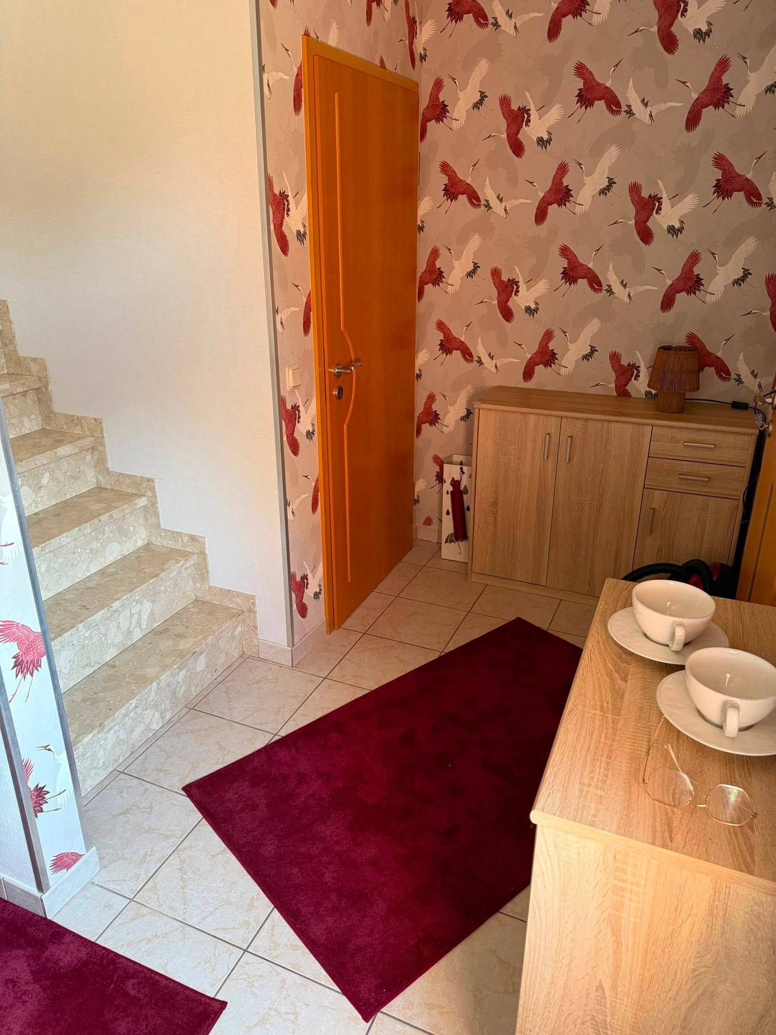 Una habitación con puerta de madera, una escalera, una zona de cocina con armarios y una alfombra roja.