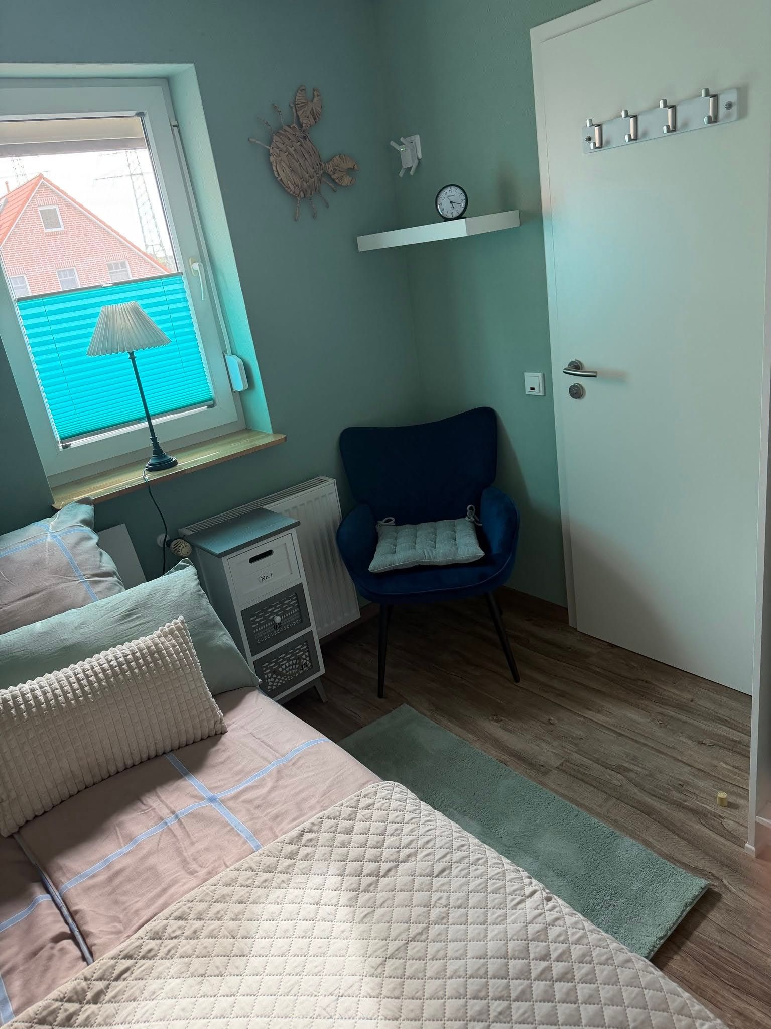 Ein Schlafzimmer mit einem Bett, einem Sessel, einem Heizkörper, einem Fenster mit Jalousien und einer Tür mit Lichtschalter.