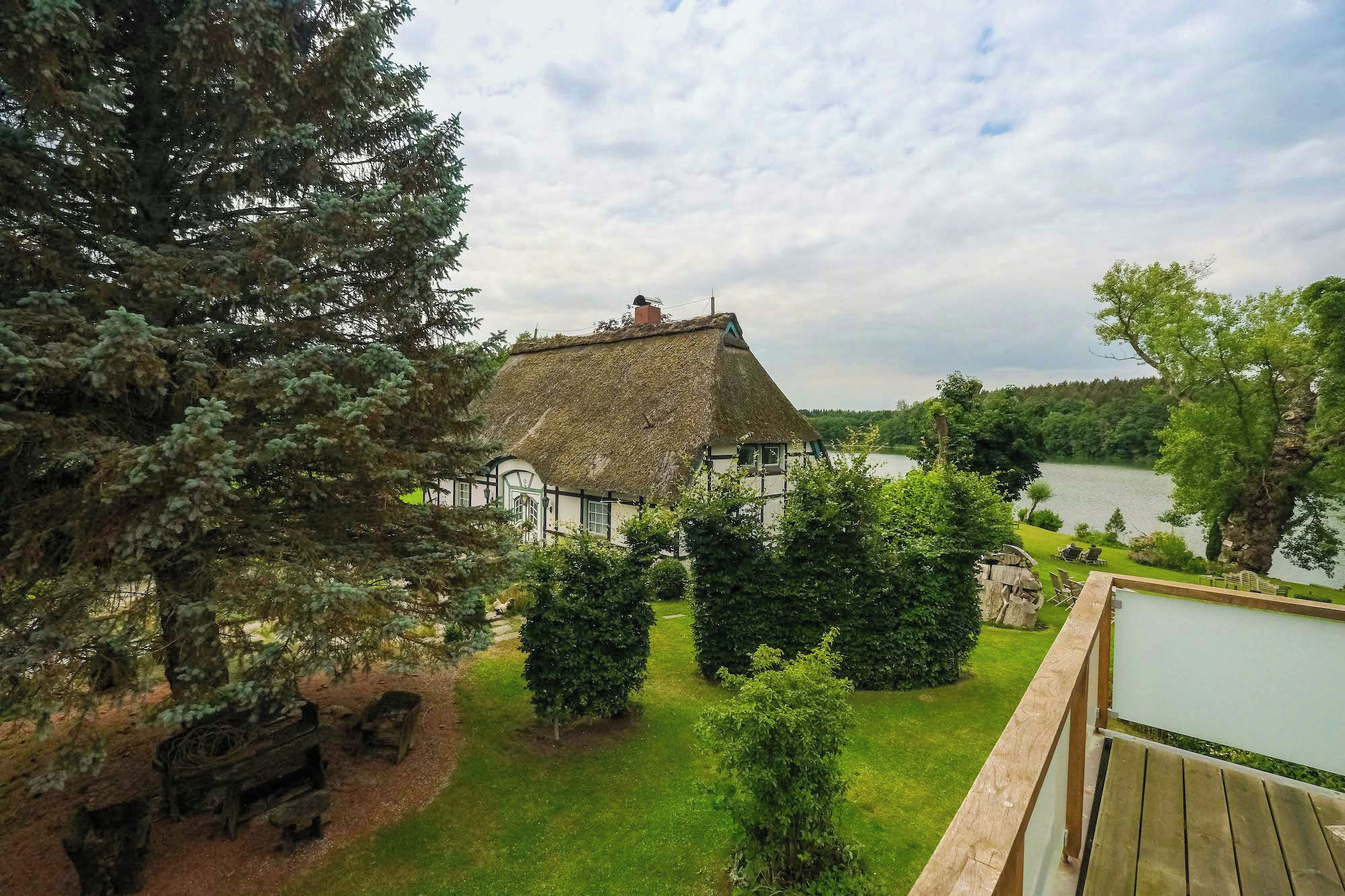 A traditional thatched-roof house with a lawn and a wooden deck overlooking a lake.