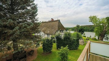 A traditional thatched-roof house with a lawn and a wooden deck overlooking a lake.