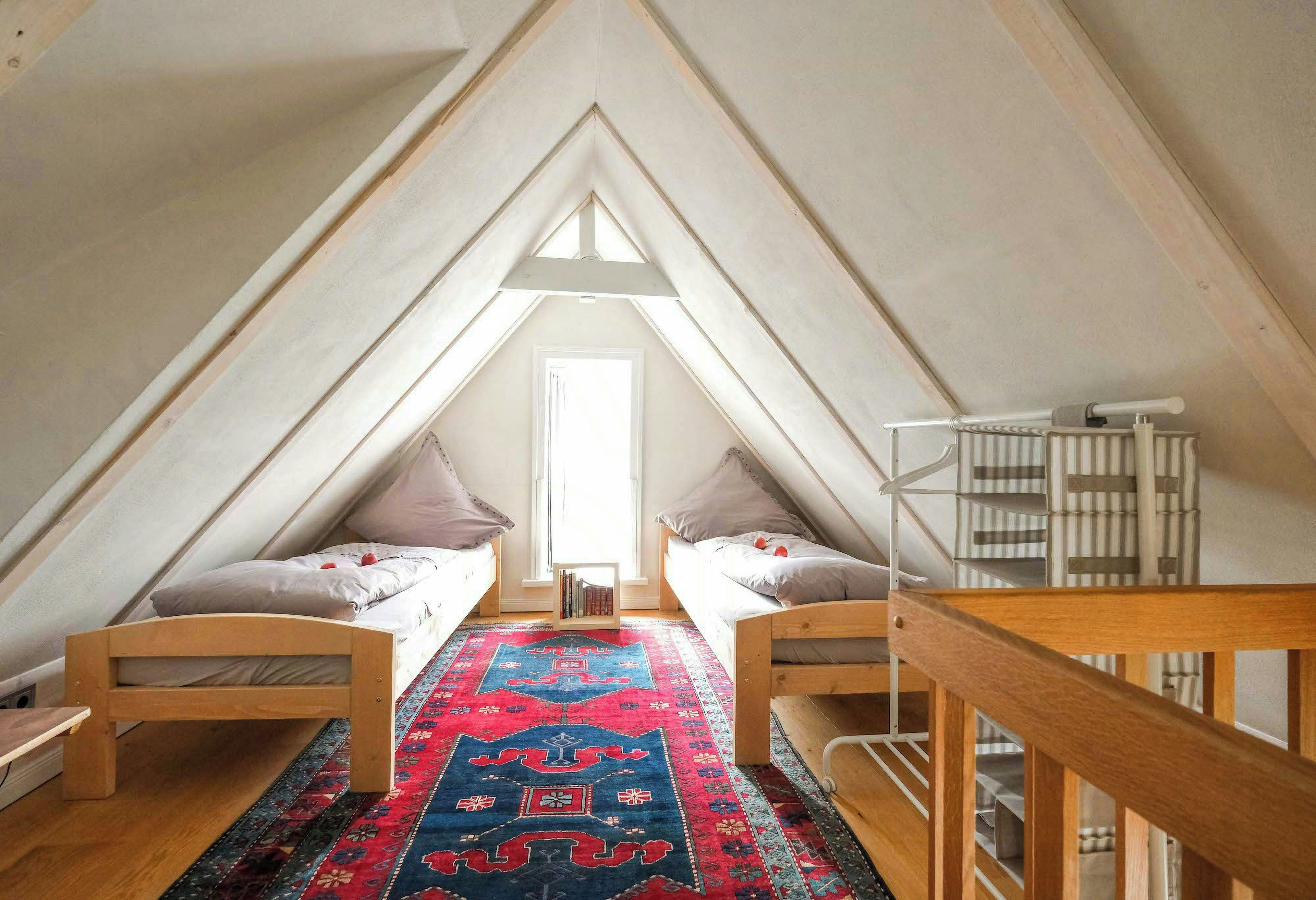 Une chambre mansardée triangulaire avec deux lits, un tapis à motifs rouges et un radiateur.