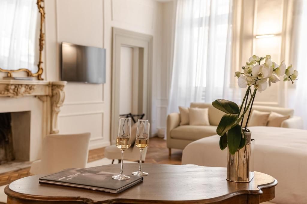 Quarto de hotel com mesa de madeira, duas taças de vinho, um vaso com flores brancas, um sofá e uma televisão na parede.