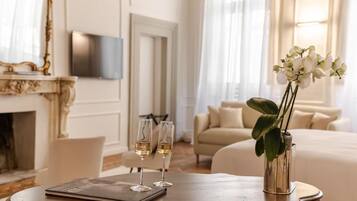 A hotel room with a wooden table, two glasses of wine, a vase with white flowers, a sofa, and a television on the wall.
