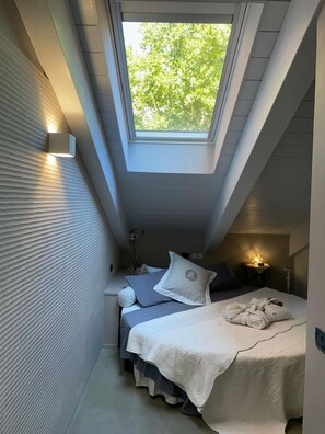 Une petite chambre à coucher, de dimensions réduites, avec un puits de lumière, un lit recouvert d’une couverture blanche et une table de chevet avec une lampe.
