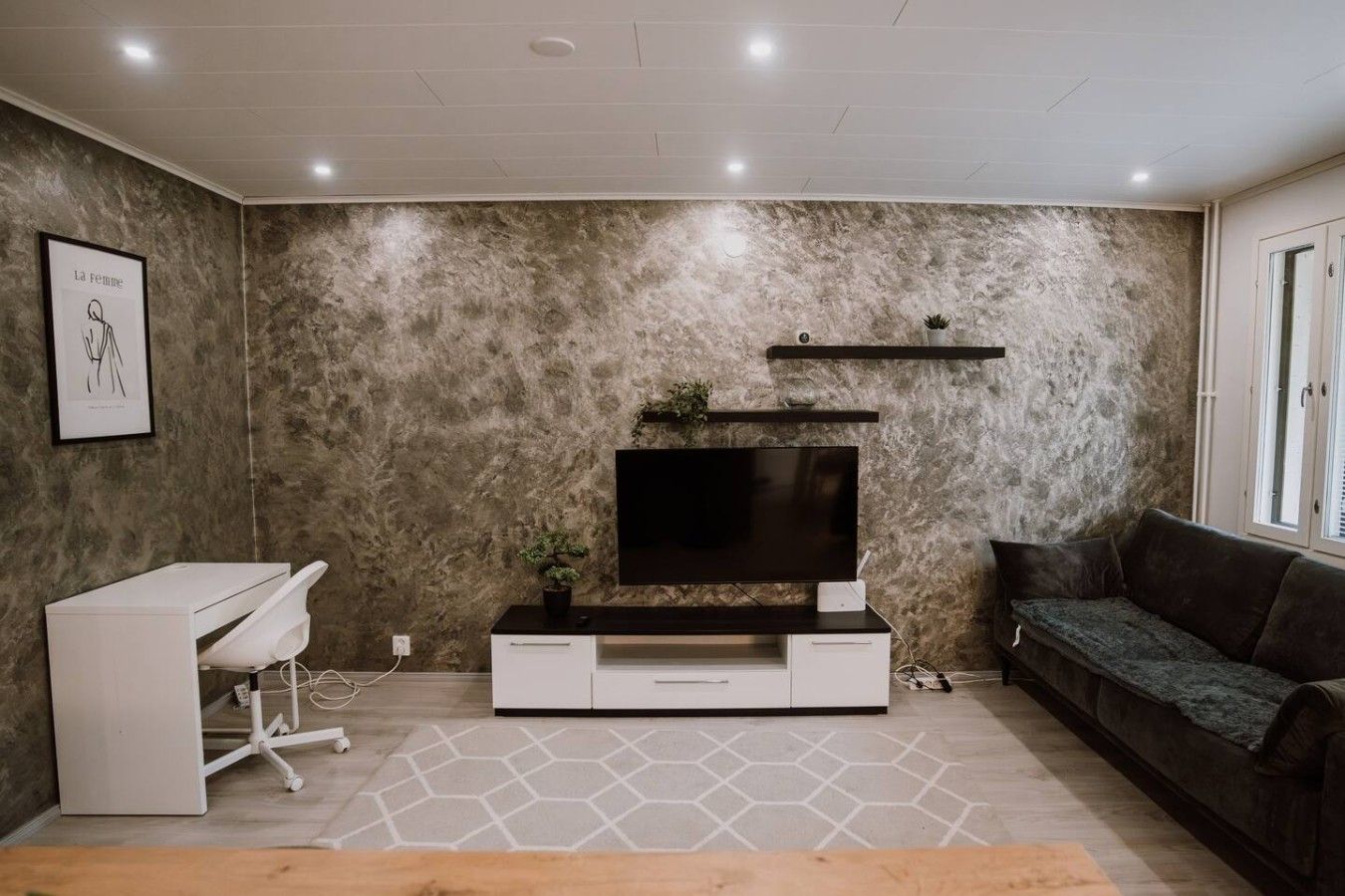 Un salon avec un téléviseur à écran plat, un bureau blanc, un canapé foncé et un tapis à motifs.