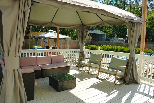 A covered outdoor seating area with cushioned benches and chairs, a glass-top table, and a wooden deck.