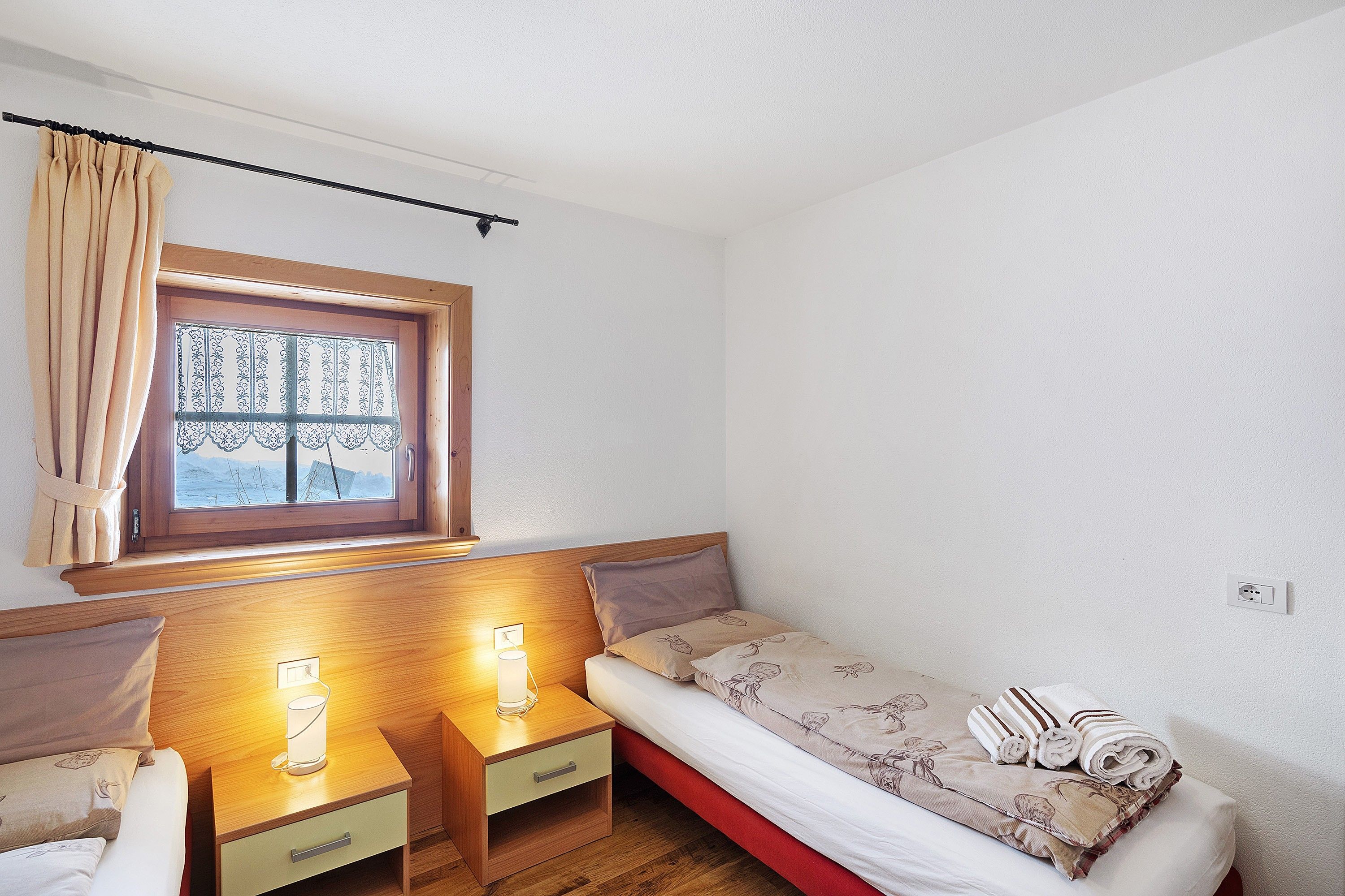 Ein kleines Zimmer mit einem Einzelbett, einem Holzkopfende, einem Nachttisch mit Lampe und einem fenster mit Vorhängen.