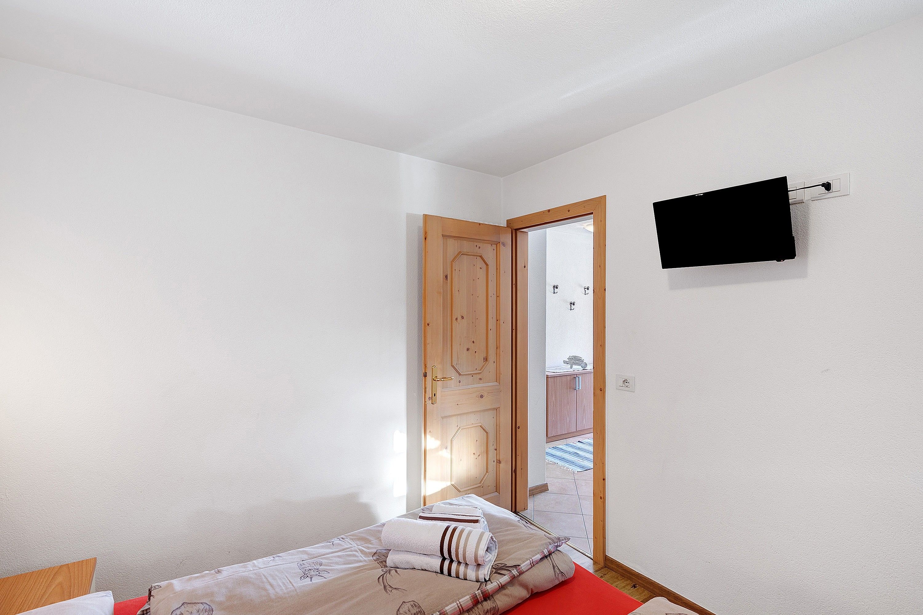Ein Zimmer mit einem Bett, einer Holztür, einem an der Wand befestigten Fernseher und Blick durch die Tür.