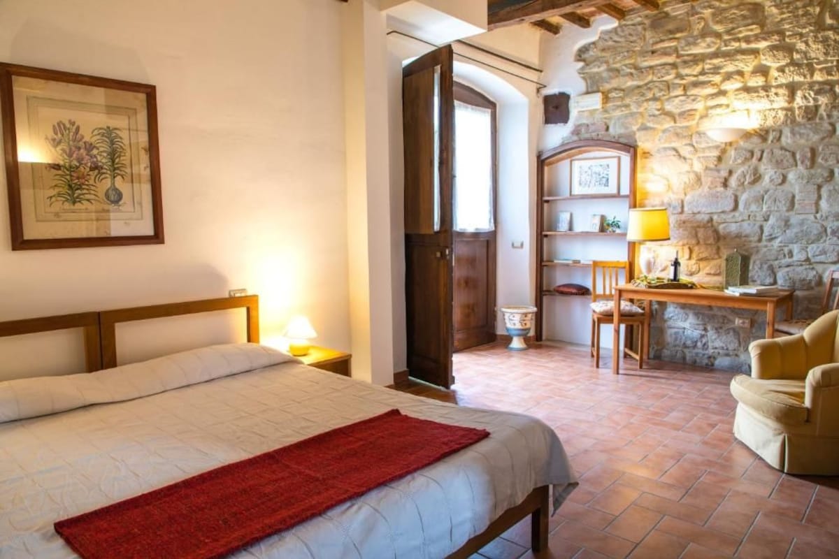 Une chambre à coucher avec un lit, un plaid rouge, une tête de lit en bois, un mur en pierre, un bureau avec une lampe et une chaise.