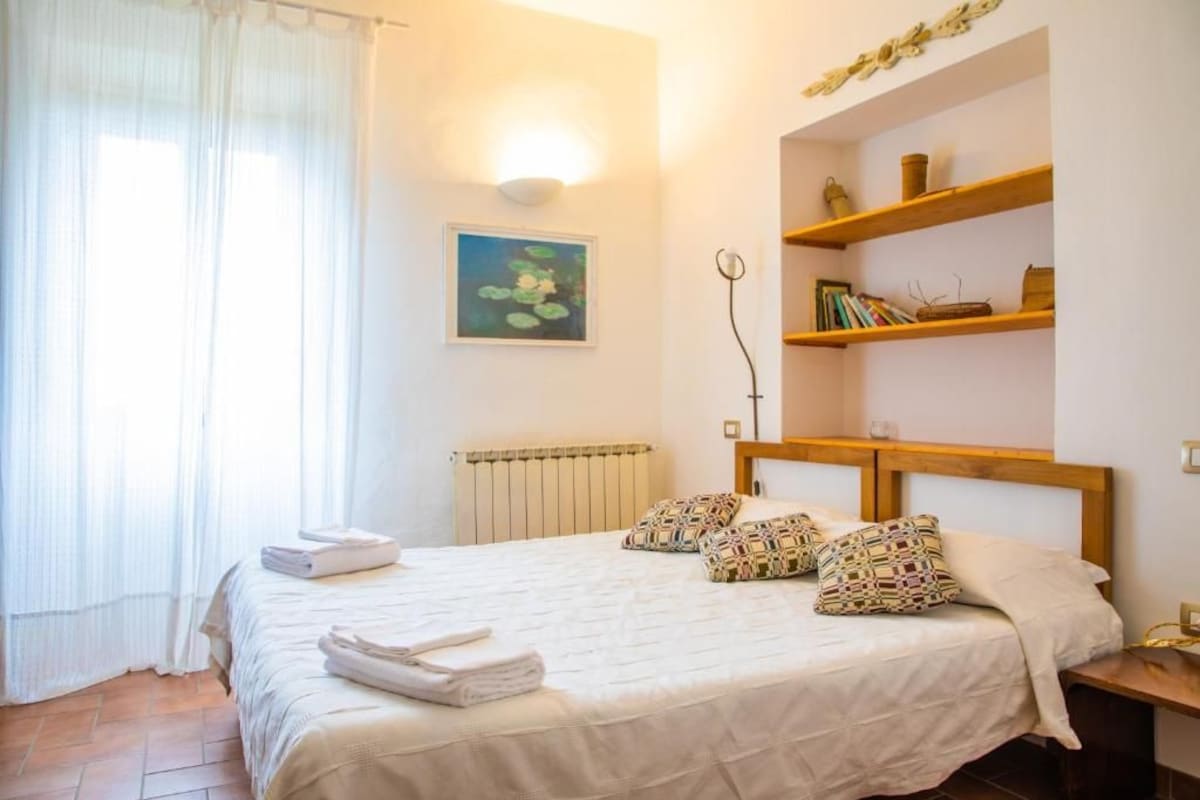 Une chambre d’hôtel avec un lit, un radiateur, un tableau accroché au mur et une étagère avec des livres et un panier.