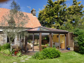 Une maison dotée d’une véranda en verre, d’une cheminée en pierre et d’un jardin où poussent différentes espèces de plantes.