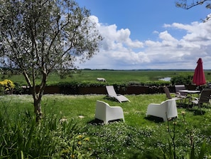Un espace extérieur verdoyant, avec des chaises longues blanches et un parasol rouge, donnant sur un champ où paissent des vaches et sur une étendue d’eau.