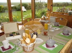 Une table mise pour le petit-déjeuner, avec couverts pour deux personnes, comprenant du pain, des boissons et un panier de fruits.