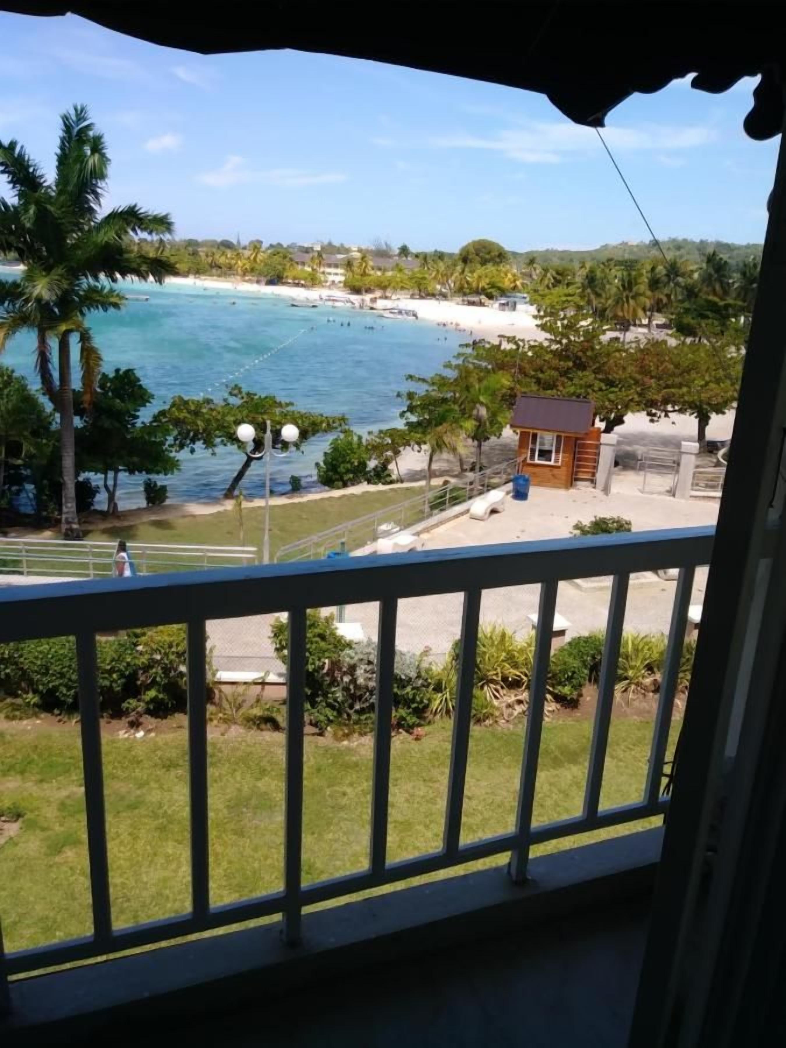 Un balcón con vistas a una playa, palmeras y un edificio pequeño.