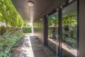 A modern building entrance with a glass door, a green exit sign, and a pathway lined with greenery. - Warsaw | Międzynarodowa 31A | Spacious Apartment | Air conditioning (Warszawa)