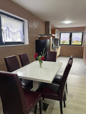 A dining area with a white table and dark chairs, a vase with flowers, and a kitchen in the background.