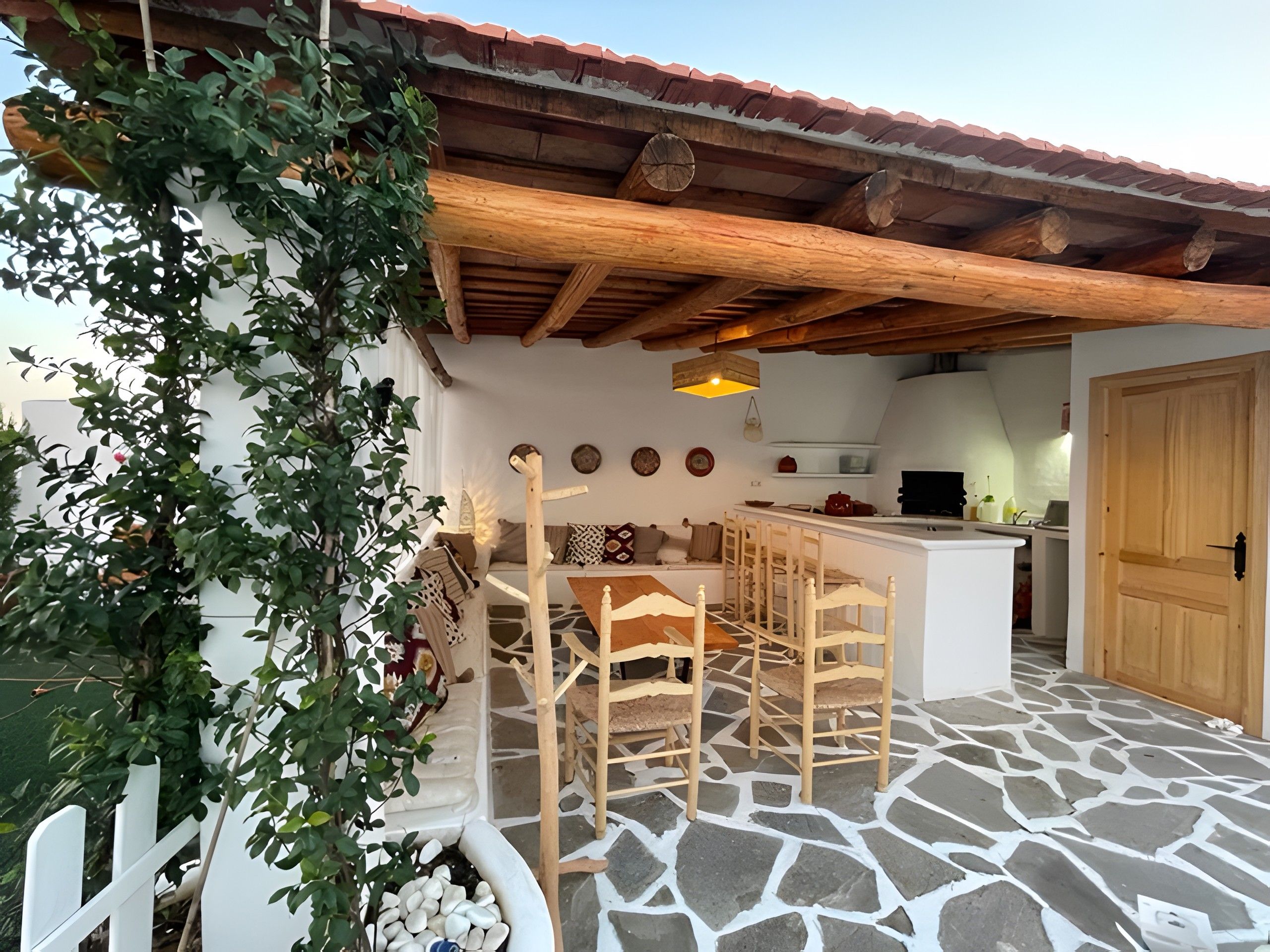 Un espace extérieur aménagé dans un style rustique, avec un sol en pierre, des meubles en bois et une cuisine en arrière-plan.