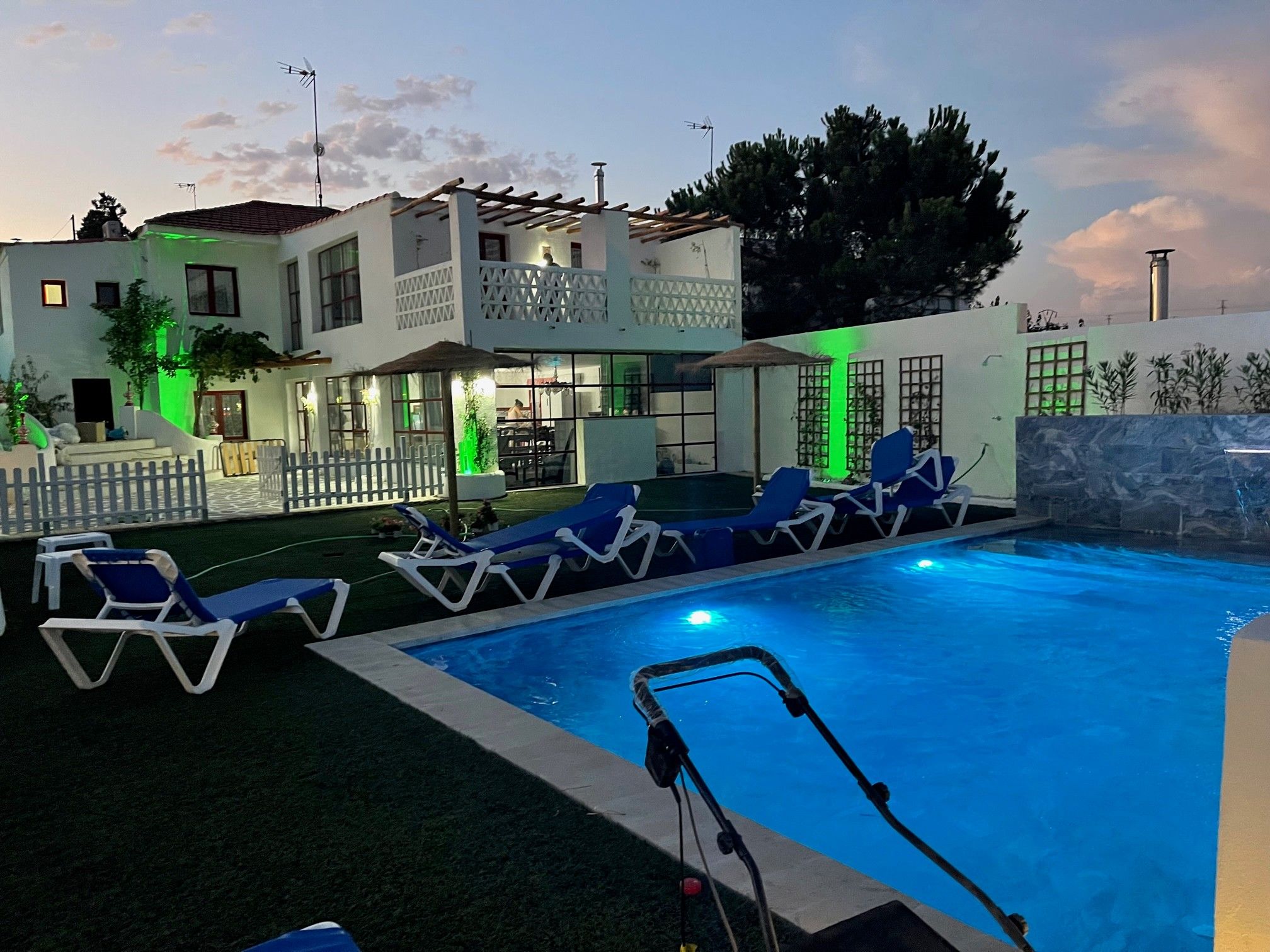 Une piscine avec des chaises longues, un espace de détente couvert et un bâtiment doté de nombreuses fenêtres et d’un balcon.