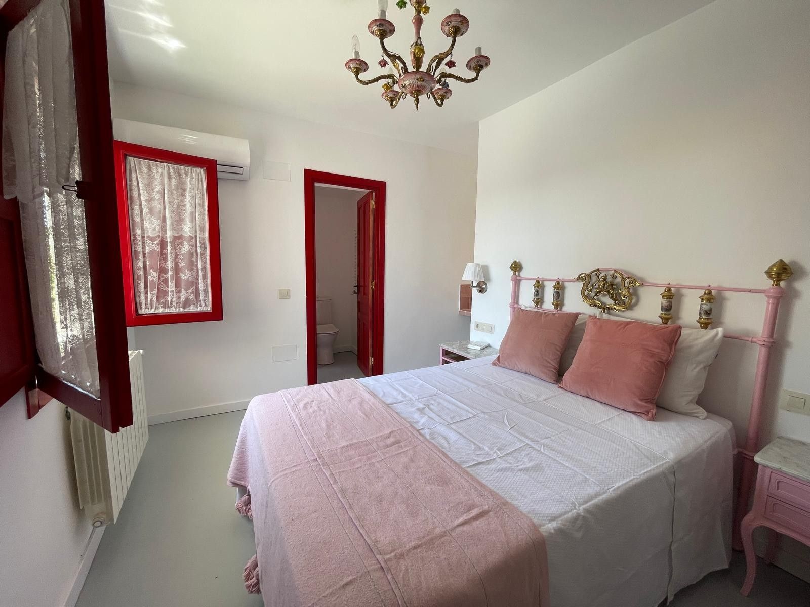 A bedroom with a bed, a nightstand, a lamp, a chandelier, and a window with curtains.