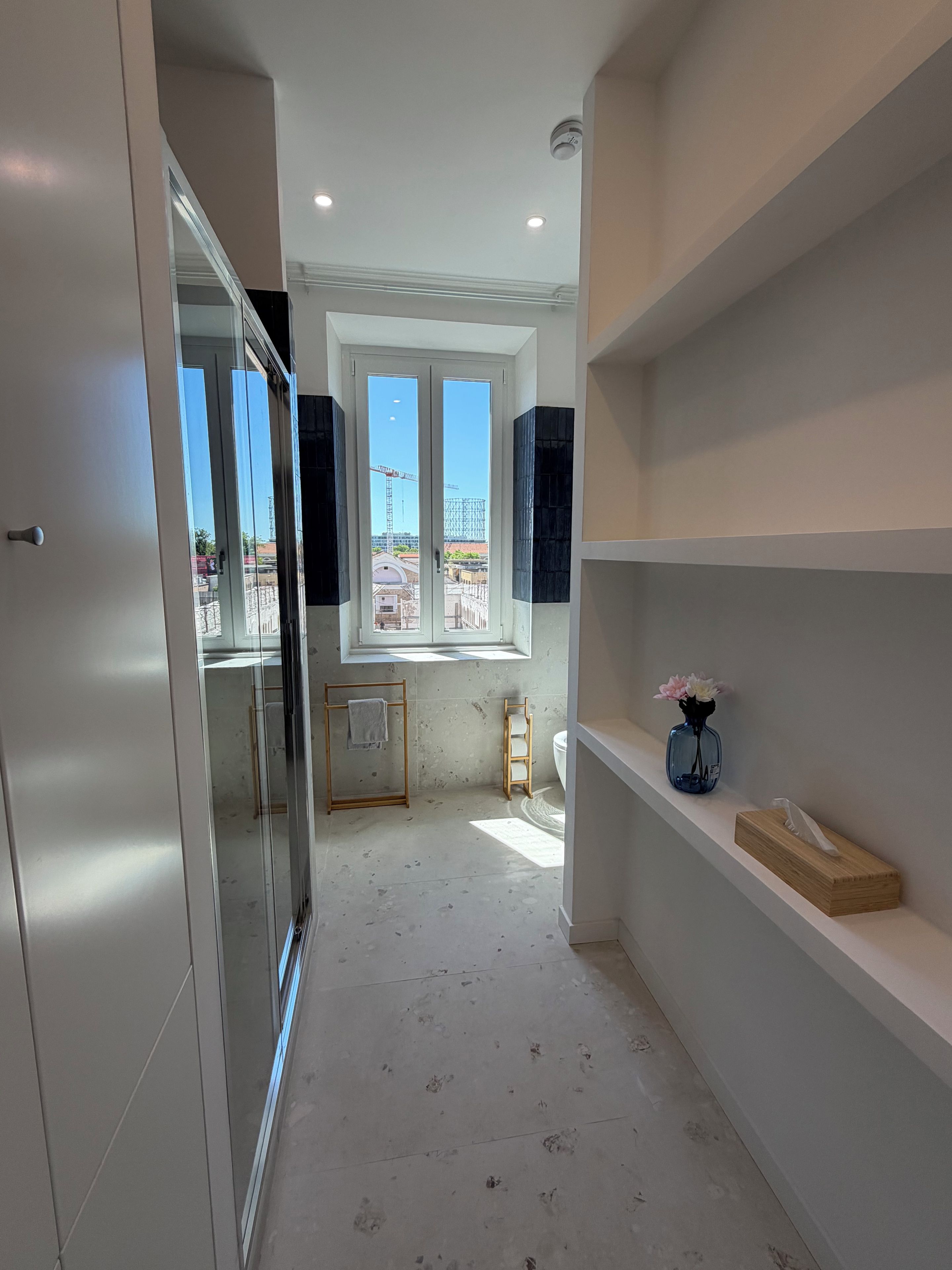 A modern bathroom with a large mirror, a window with a view of construction, and a shelf with a tissue box.