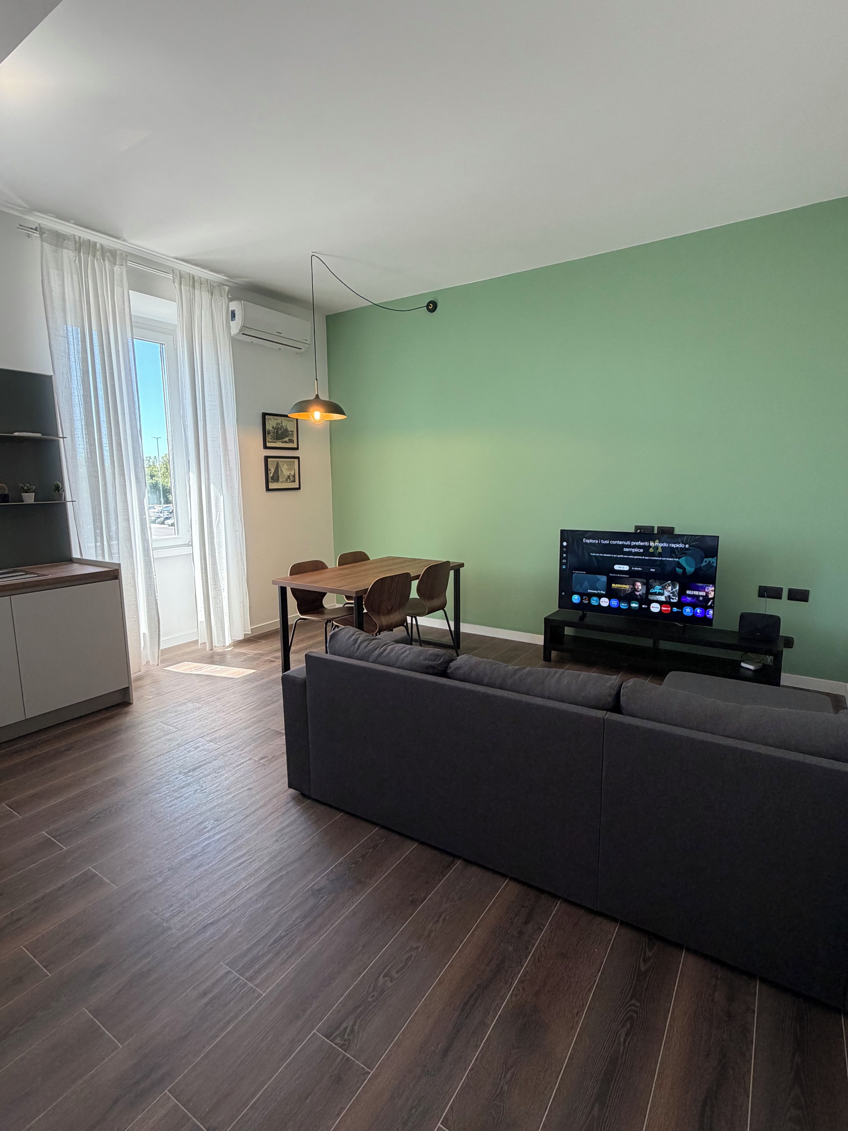 A modern living room with a sofa, a dining table with chairs, a television, and a kitchen area with a window.