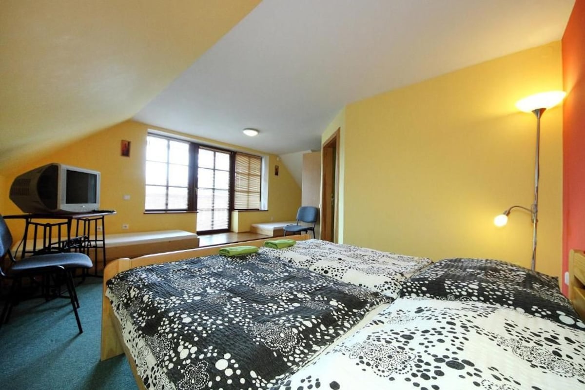Quarto com cama, escrivaninha, cadeira, televisão e janela com persianas.