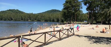 A beach with a wooden fence, people sunbathing, and a forested hill in the background.