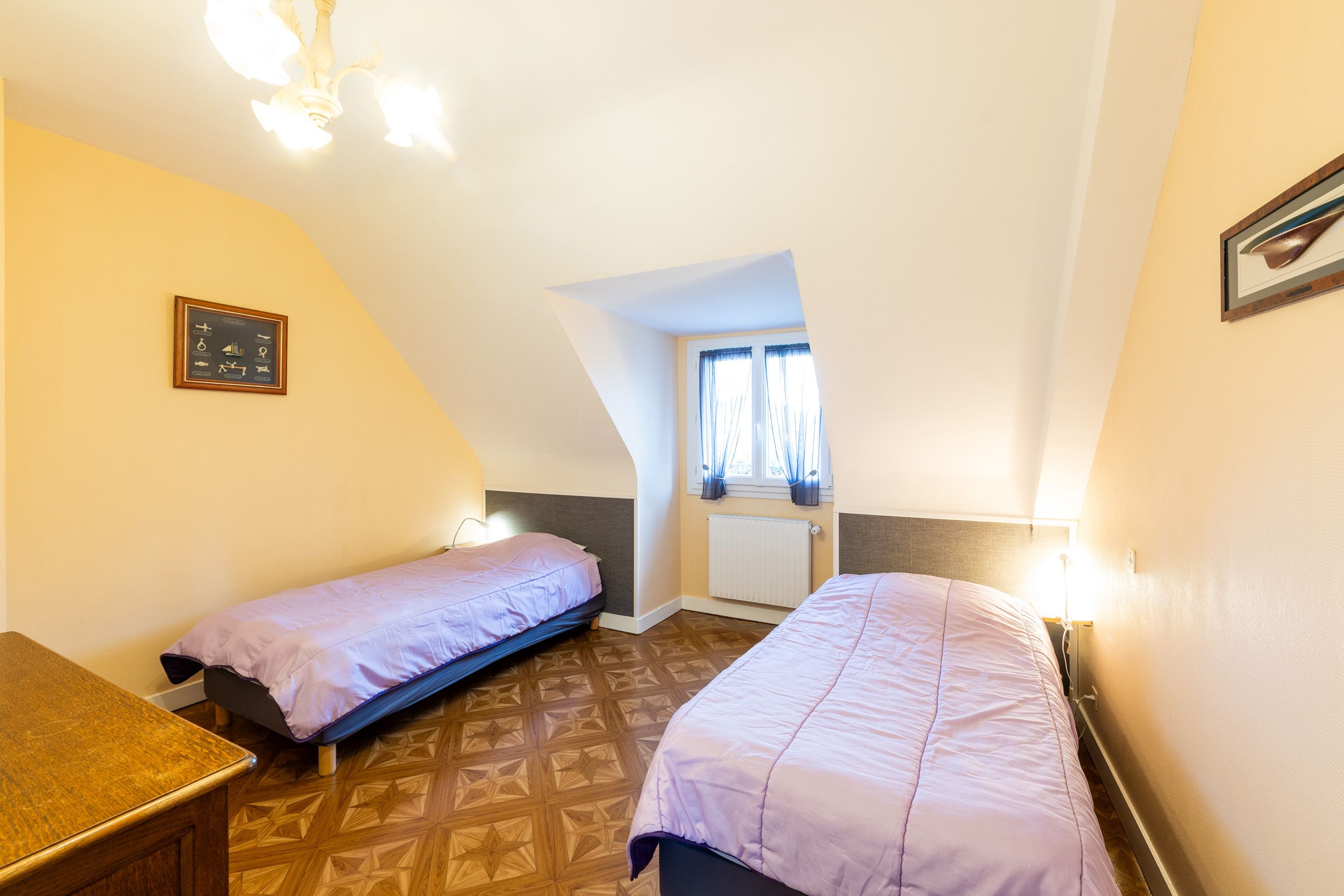 Een kleine, gezellige kamer met twee eenpersoonsbedden, een houten vloer, een inlijsting foto aan de muur en een raam met gordijnen.