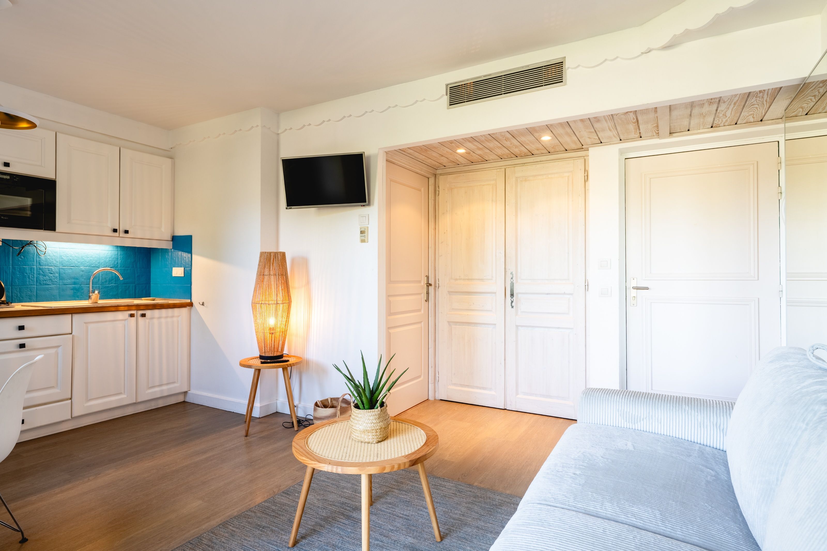 Un salon moderne comprenant un canapé, une petite table ronde avec une lampe, et un coin cuisine avec des placards et un téléviseur.