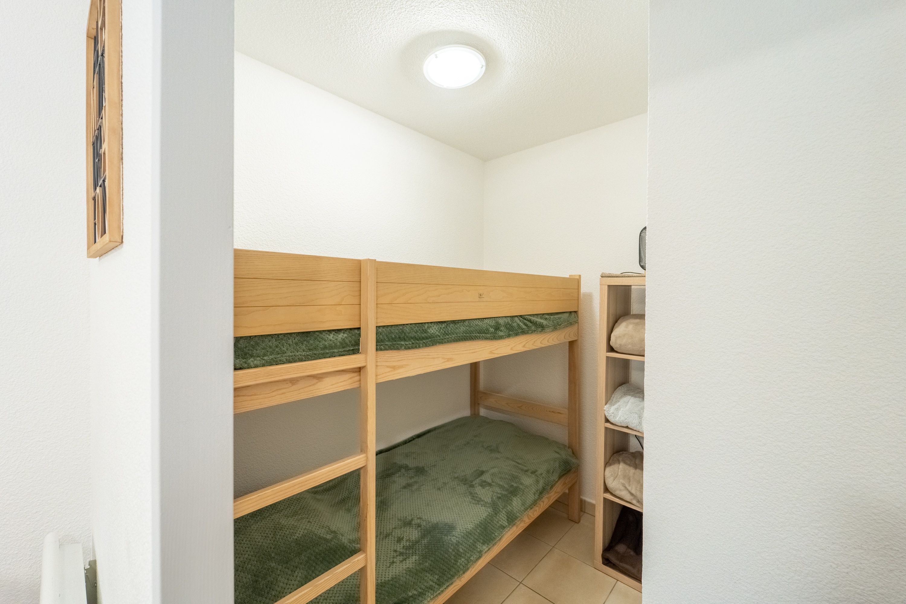A bunk bed with a green mattress and a wooden headboard, a small shelf with a cushion, and a white wall with a single light fixture.